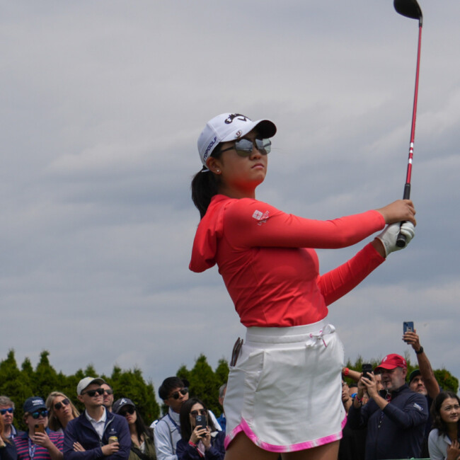 Woman golfing with a white skirt and red shirt with hat and sunglasses and a crowd in the background