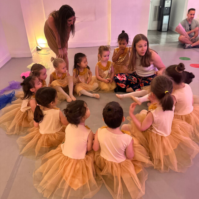 Dancers in yellow tutus and white shirts sit on the ground in a circle with one adult teacher sitting with them and one adult teacher standing behind them