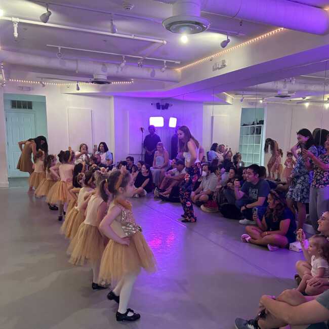 A group of young dancers in yellow tutus performs in front of a crowd of their parents in the AXIS studio