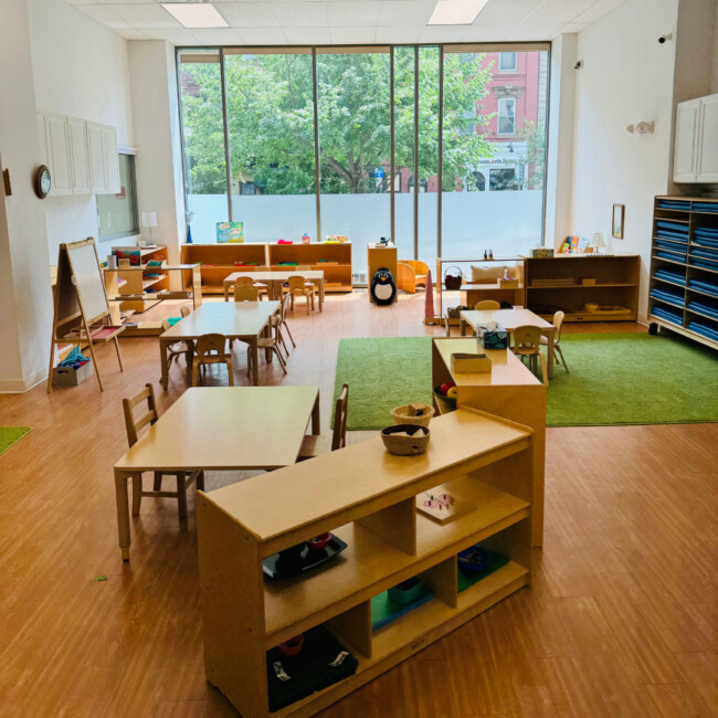 Classroom view with large windows, green rug, wood shelves, table, chairs, and art easel