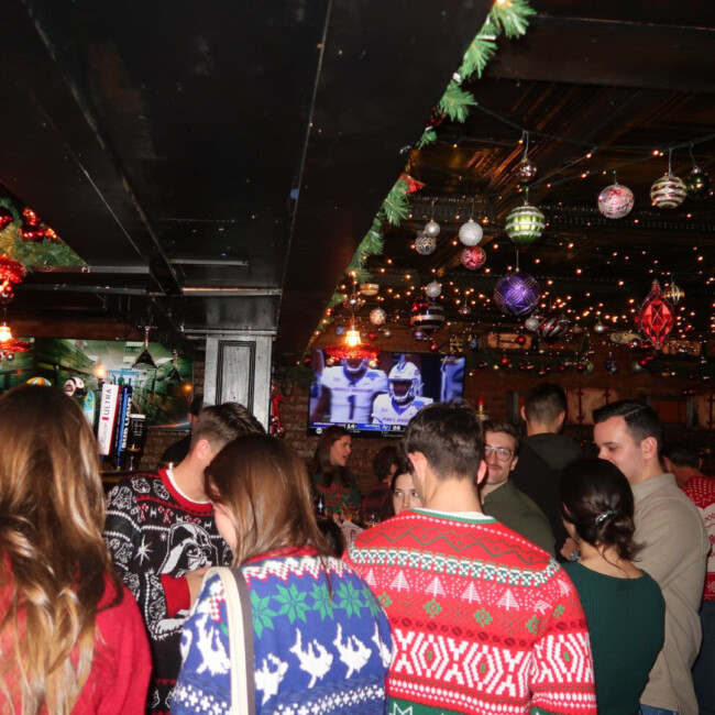 A group of people wearing Christmas sweaters in The Waiting Room with football on the TV in background